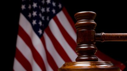 A wooden table features a judge's gavel, positioned in front of an American flag, representing the legal system and governance.